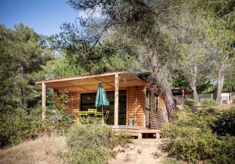 Cabaña de madera con terraza, mobiliario de jardín y sombrilla en el bosque de Huttopia La Forêt de Janas.