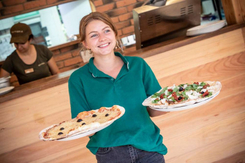 Camarera sonriente con polo verde sirviendo dos pizzas en el restaurante de Huttopia La Forêt de Janas, Francia.