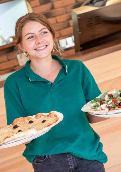Camarera sonriente con polo verde sirviendo dos pizzas en el restaurante de Huttopia La Forêt de Janas, Francia.