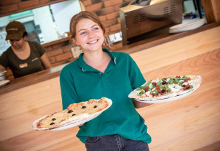 Camarera sonriente con polo verde sirviendo dos pizzas en el restaurante de Huttopia La Forêt de Janas, Francia.