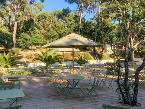 Zona de cafetería al aire libre con mesas, sillas y sombrilla, rodeada de árboles en un parque vacacional de Provenza, Francia.