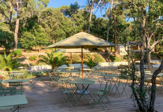 Buitenterras met tafels, stoelen en parasol, omringd door bomen in een vakantiepark in de Provence, Frankrijk.