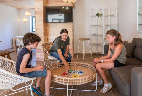 Kinderen en een volwassene spelen een bordspel in de lounge van Huttopia La Forêt de Janas vakantiepark.