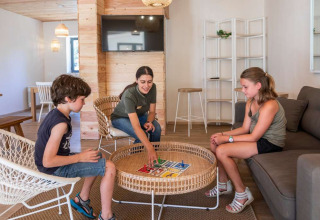 Kinderen en een volwassene spelen een bordspel in de lounge van Huttopia La Forêt de Janas vakantiepark.