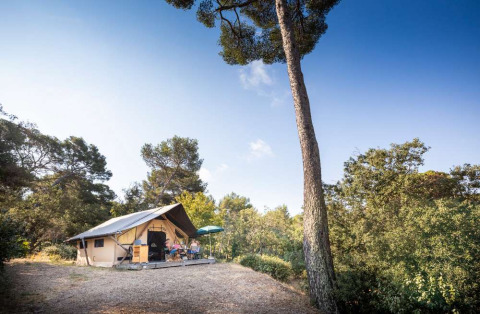 Glamping tent and people relaxing on a terrace at Huttopia La Forêt de Janas, surrounded by pine trees.