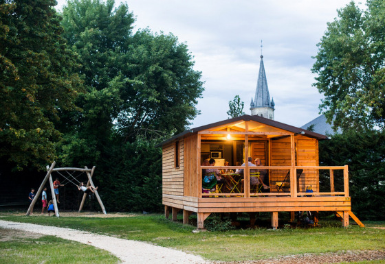 Cabaña de madera Chalet Evasion con personas en el porche y niños jugando en columpios cerca al atardecer.