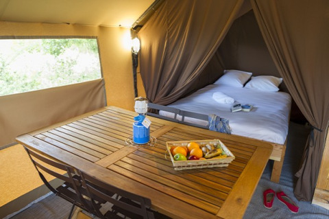 Interior de tienda safari con cama doble, mesa de madera, canasta de frutas y luz acogedora.
