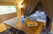 Interior de tienda safari con cama doble, mesa de madera, canasta de frutas y luz acogedora.