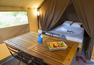 Interior de tienda safari con cama doble, mesa de madera, canasta de frutas y luz acogedora.