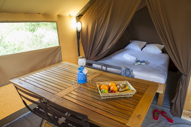 Intérieur de tente safari confortable avec lit double, table en bois, panier de fruits et ambiance chaleureuse.