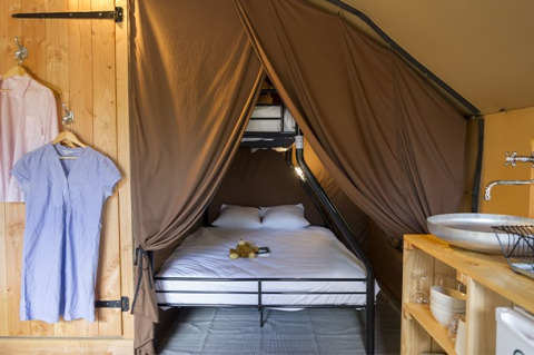 Interior de una tienda safari con cama matrimonial, armario abierto con ropa y lavabo rústico de madera.