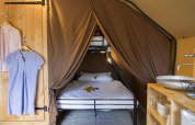 Interior de una tienda safari con cama matrimonial, armario abierto con ropa y lavabo rústico de madera.