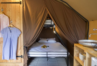 Interior de una tienda safari con cama matrimonial, armario abierto con ropa y lavabo rústico de madera.