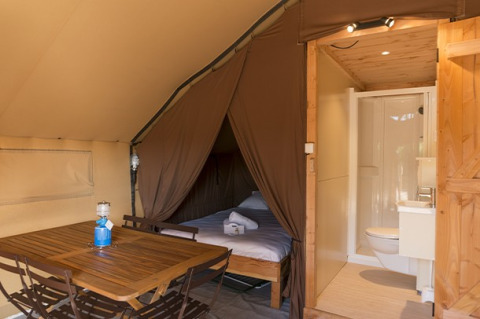 Innenraum eines Safarizeltes mit Holzmöbeln, Schlafbereich mit Bett und eigenem Bad mit WC und Dusche.