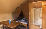 Interno di una tenda safari con mobili in legno, letto nella zona notte, e bagno privato con doccia.