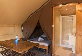 Innenraum eines Safarizeltes mit Holzmöbeln, Schlafbereich mit Bett und eigenem Bad mit WC und Dusche.