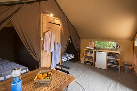 Interior view of a safari tent featuring a dining table, kitchenette, beds, and clothes hanging on hooks.