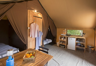 Binnenzicht van een safaritent met eettafel, keukentje, bedden en kleding aan haken aan de muur.