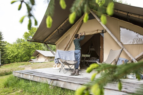 Persoon opent een safaritent op een houten platform, glamping tussen het groen in een bosrijke setting.