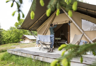 Persoon opent een safaritent op een houten platform, glamping tussen het groen in een bosrijke setting.