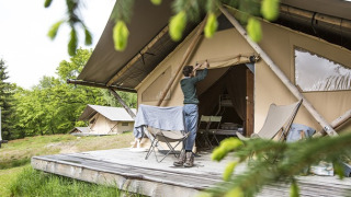 Une personne ouvre un safari-tente sur une terrasse en bois, glamping dans la forêt entourée de verdure.