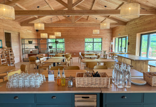 Salle à manger spacieuse en bois avec verres, carafes et vue sur la verdure par les fenêtres panoramiques.