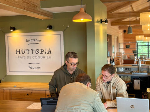 Staff at Huttopia Pays de Condrieu assist a guest at check-in in a cozy, wood-accented reception area in France.