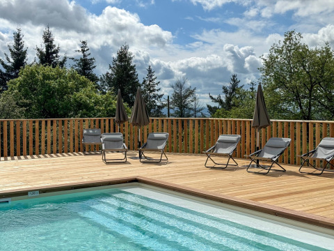Udendørs swimmingpool med liggestole og parasoller på træterrasse, omgivet af træer og blå himmel.