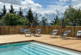 Udendørs swimmingpool med liggestole og parasoller på træterrasse, omgivet af træer og blå himmel.
