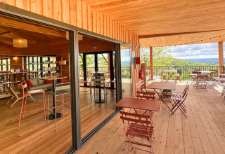 Holzterrasse und Innenraum im Village Huttopia Pays de Condrieu, Frankreich, mit Tischen und Panoramablick.