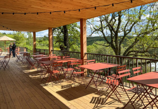Udendørs terrasse med røde borde og stole, træloft og udsigt over træer i Village Huttopia Pays de Condrieu.