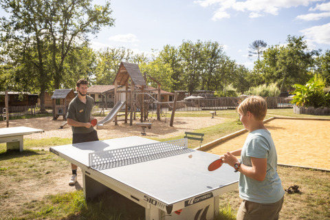 Dos personas juegan al ping-pong al aire libre junto a un parque infantil en Village Huttopia Pays de Condrieu.