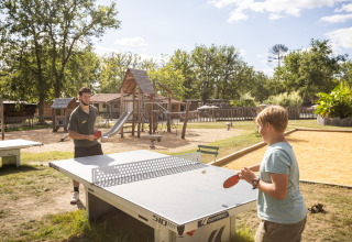 Dos personas juegan al ping-pong al aire libre junto a un parque infantil en Village Huttopia Pays de Condrieu.