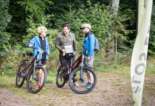Trois personnes discutent avec des vélos au Village Huttopia Pays de Condrieu dans une forêt verdoyante.