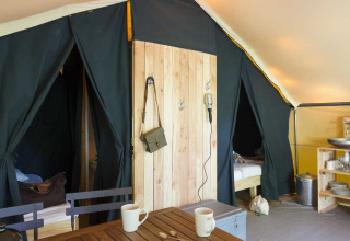Binnenzicht van een safaritent met bedden, een keukentje en een houten tafel met kopjes en servies.