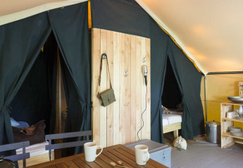 Intérieur d'une tente safari avec lits, coin cuisine, table en bois et mugs dans une ambiance chaleureuse.