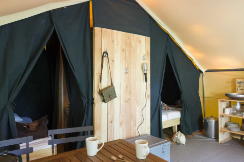 Intérieur d'une tente safari avec lits, coin cuisine, table en bois et mugs dans une ambiance chaleureuse.