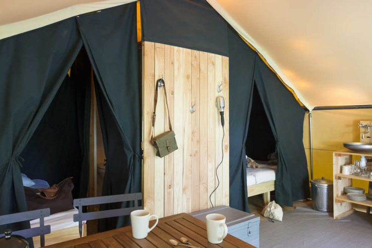 Vista interior de una tienda safari con camas, una pequeña cocina y una mesa de madera con tazas.