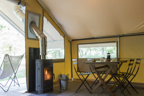 Intérieur d'une tente safari avec poêle à bois, table à manger, chaises pliantes et lumière naturelle.