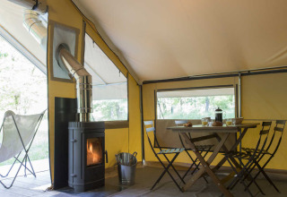 Intérieur d'une tente safari avec poêle à bois, table à manger, chaises pliantes et lumière naturelle.