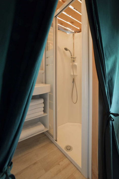 Shower cabin and towel shelves inside a safari tent with wood flooring and green curtains pulled aside.