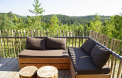 Coin salon extérieur sur la terrasse d’une tente safari avec meubles en bois et vue sur la forêt verdoyante.