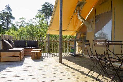 Buitenterras bij een safaritent met houten loungeset, eetset, boomstamtafeltjes en uitzicht op het bos.