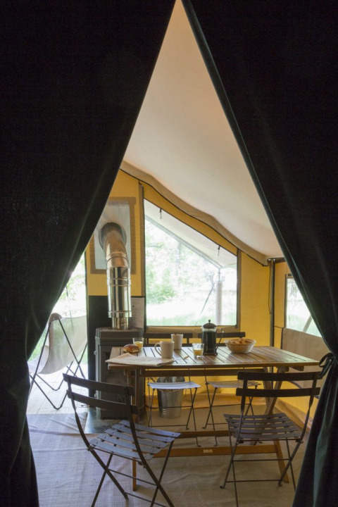 Binnenzicht van een safaritent met donker gordijn, houten tafel, stoelen en een houtkachel bij raam.