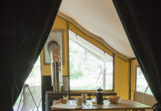 Binnenzicht van een safaritent met donker gordijn, houten tafel, stoelen en een houtkachel bij raam.