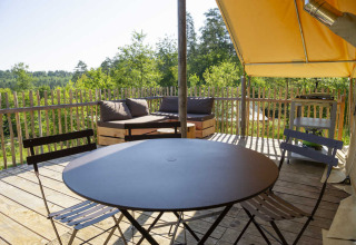 Terras van safaritent met ronde tafel, stoelen, houten bank en hekwerk in een groene omgeving.
