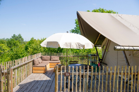 Terraza de madera con sofá, mesa, sombrilla y tienda safari, rodeada de árboles verdes y cielo despejado.