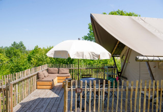 Wooden deck with a sofa, table, umbrella, and safari tent, surrounded by lush green trees in sunlight.