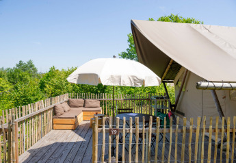 Deck in legno con divano, tavolo, ombrellone e tenda safari, circondato da alberi verdi e cielo sereno.