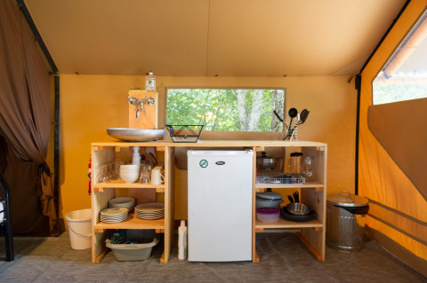 Cocina en tienda safari con nevera pequeña, fregadero, platos, utensilios y estanterías de madera.